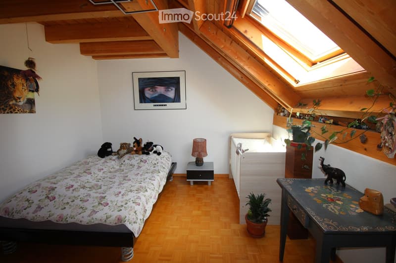 Cozy attic bedroom with wooden slanted ceilings, skylights, a single bed with floral bedding, nightstand with lamp, small table with decorative items, and potted plants.
