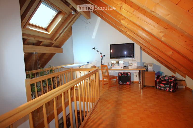 Attic room with wooden beams, skylight windows, wooden floor, and a desk area with a sewing machine, chair, and television mounted on the wall.