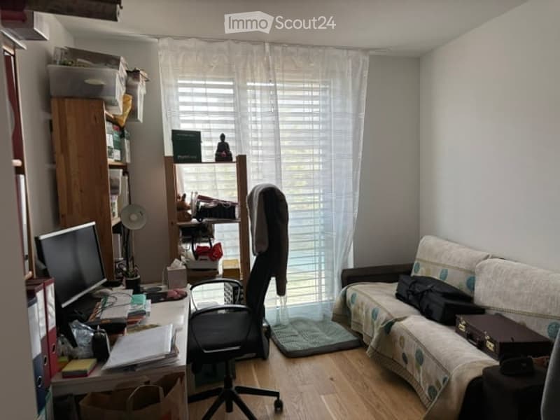 A small, furnished room with a wooden floor, a sofa with a patterned cover, a desk with a computer, a chair, and a shelf with books and other items. A window with white curtains allows natural light to enter.
