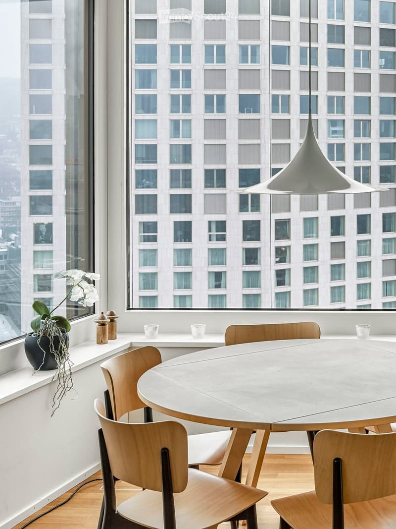 Dining area with a round table, wooden chairs, a white wall, a window with a view of the city, a white lamp, and a potted plant on the window sill
