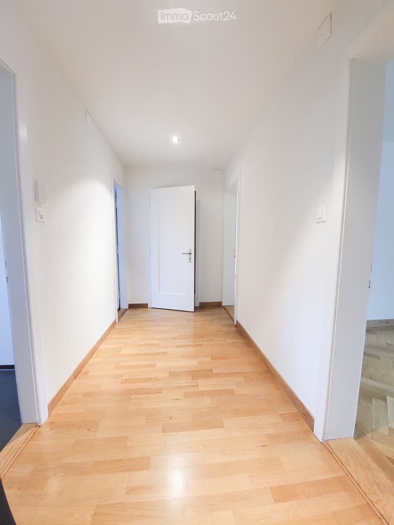 A clean, well-lit hallway with light wooden flooring and white walls. Features include a central door and several wall-mounted light switches.