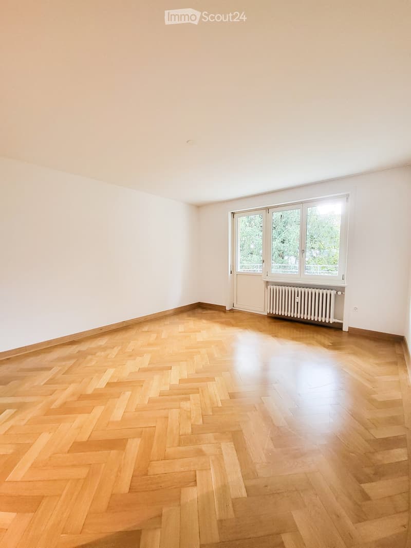 Empty room with light wooden herringbone flooring, white walls, large windows, and a radiator under the windows.