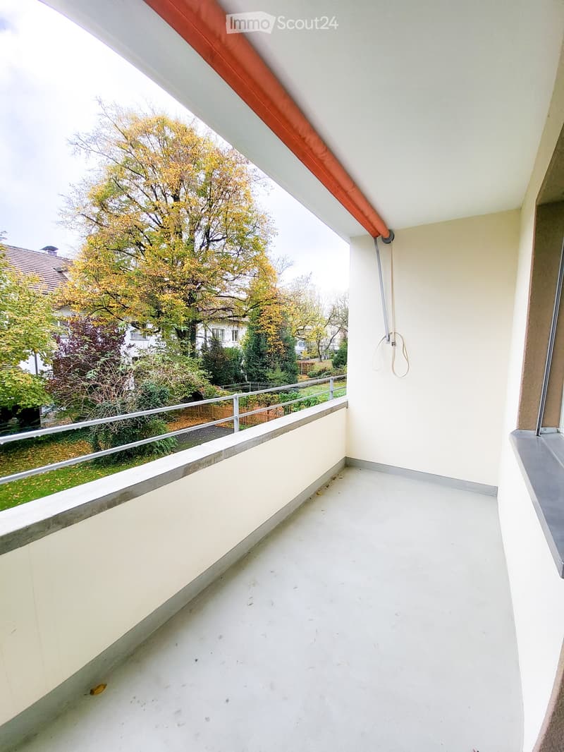 Balcony with white walls, metal railing, and a view of trees and neighboring buildings.