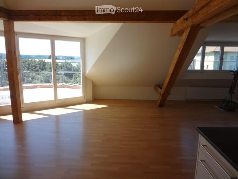 Bright, empty attic room with wooden floors, large windows, and exposed wooden beams. Features a balcony area with outdoor view.