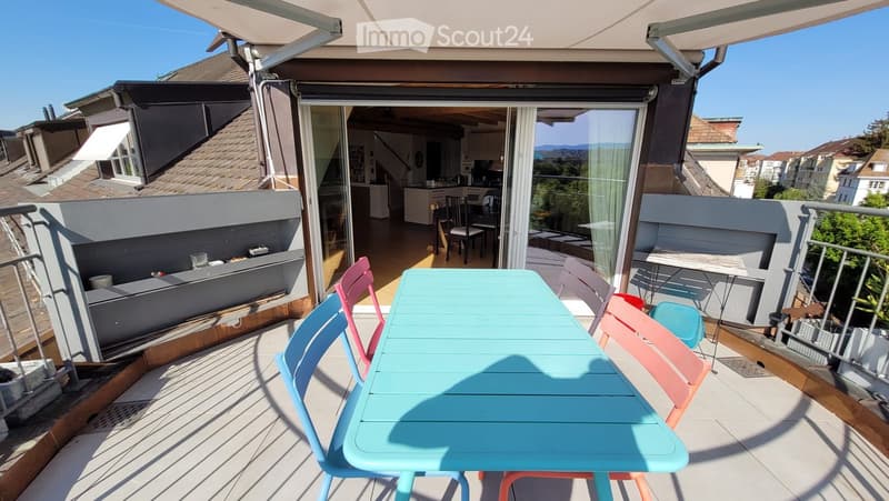 Balcony with a turquoise table and colorful chairs, overlooking a cityscape with visible rooftops and greenery.