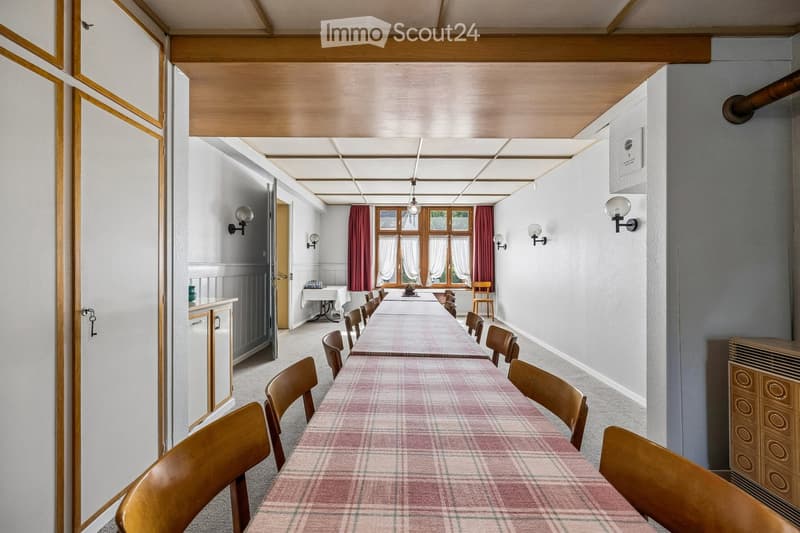 A long, narrow dining area with a long table and multiple chairs, wooden cabinetry, and wall-mounted lamps.