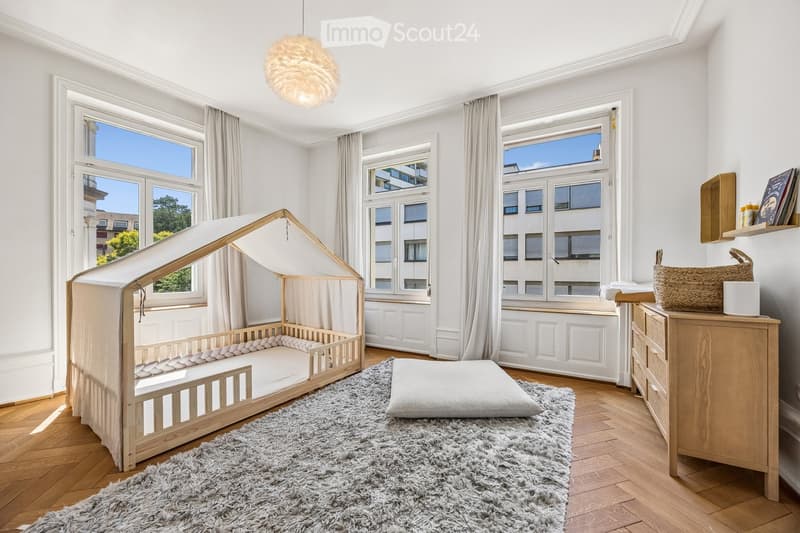 Bright, modern bedroom with wooden bed, nightstand, rug, and shelves. Large windows with curtains provide natural light.
