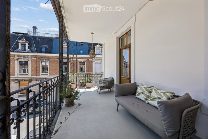 Balcony with a grey sofa, patterned cushions, a wicker chair, and several potted plants. The balcony has a metal railing and overlooks a cityscape with classic buildings.