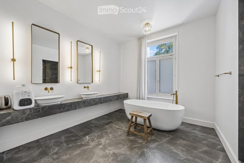 Modern bathroom with a freestanding bathtub, marble countertops, two sinks, large mirrors, and a window with white curtains.