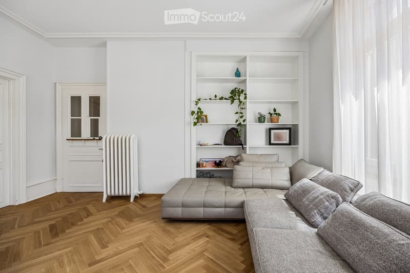 Modern living room with a grey sectional sofa, white built-in bookshelf, wooden herringbone flooring, and a radiator near the door.