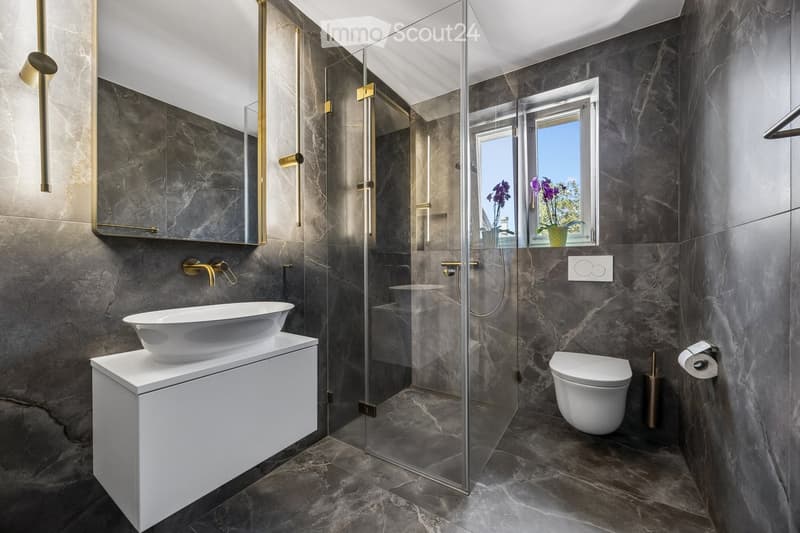 Modern bathroom with dark marble walls and floors, white sink, toilet, and glass shower enclosure. Features include a large mirror, gold fixtures, and a window with a view of purple flowers.