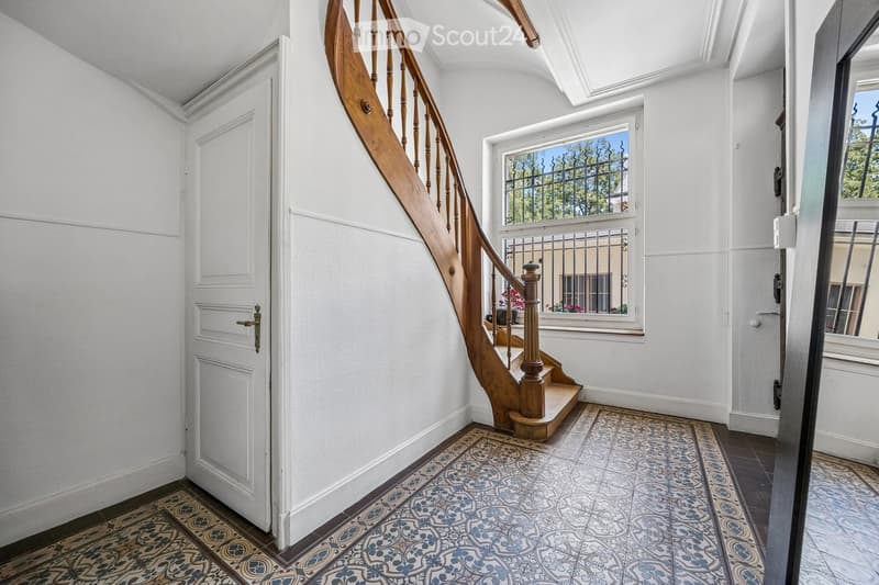Elegant hallway with a wooden staircase, decorative tiles, and white walls. Features a window with metal bars and a door on the left.