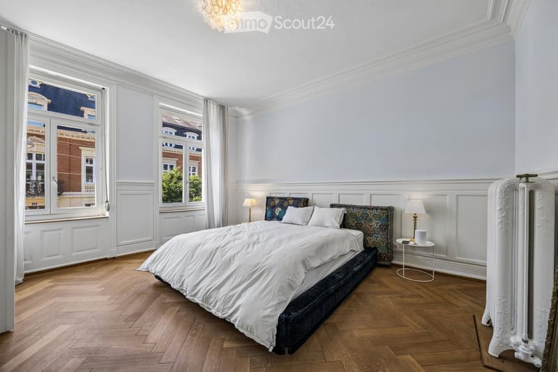Elegant bedroom with a large bed, patterned headboard, nightstands, lamps, and large windows with curtains. Wooden flooring and white walls.