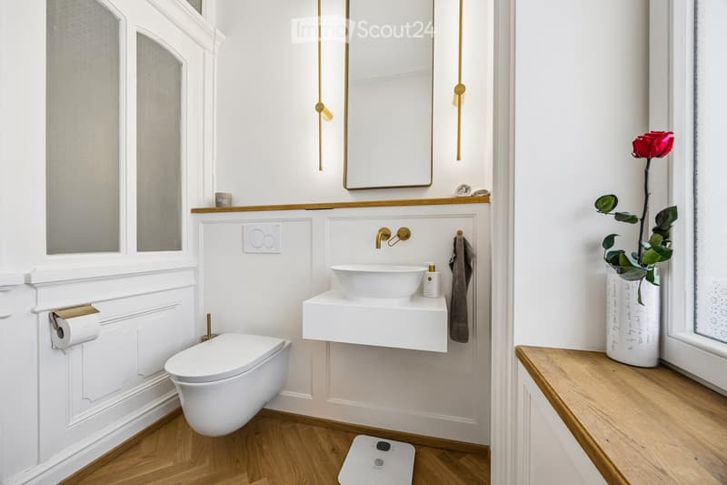 Modern bathroom with white walls, wooden flooring, a toilet, a floating sink, a mirror, a towel, a vase with a red rose, and a scale on the floor.