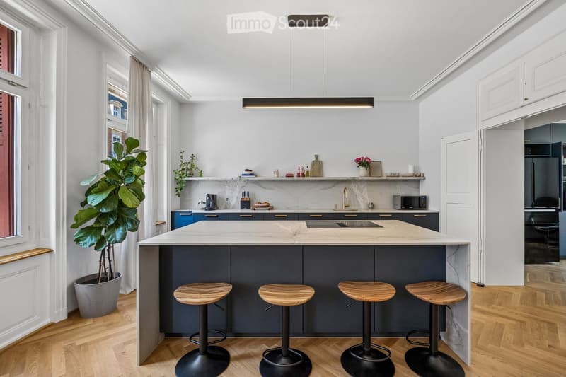 Modern kitchen with a central island, four wooden stools, and various appliances including a microwave and refrigerator. The island features a marble countertop, and there is a potted plant near the window.
