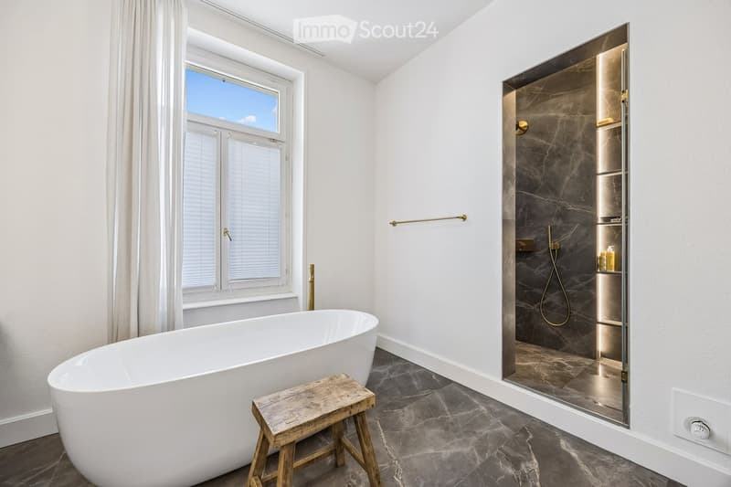 Modern bathroom with a freestanding bathtub, wooden stool, large window with blinds, and a walk-in shower with marble tiles and built-in storage.