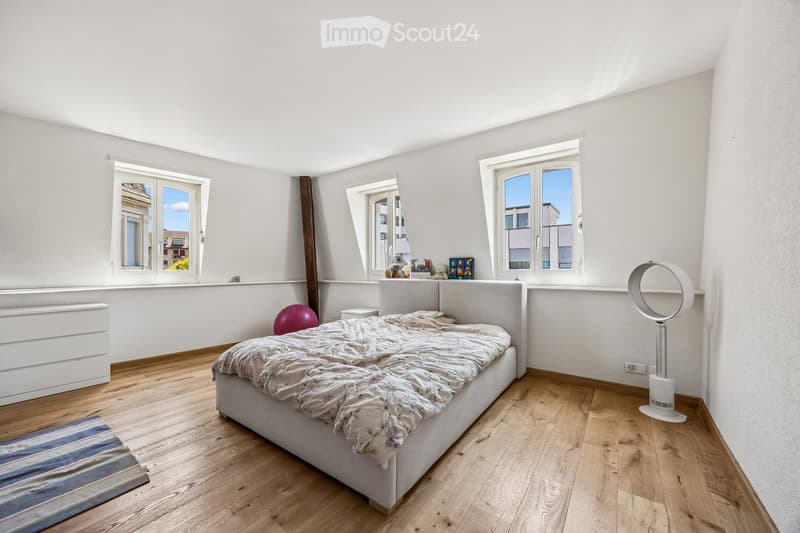 Bright bedroom with wooden flooring, large windows, a bed with a patterned duvet, a nightstand, a fan, and a colorful ball on the floor.