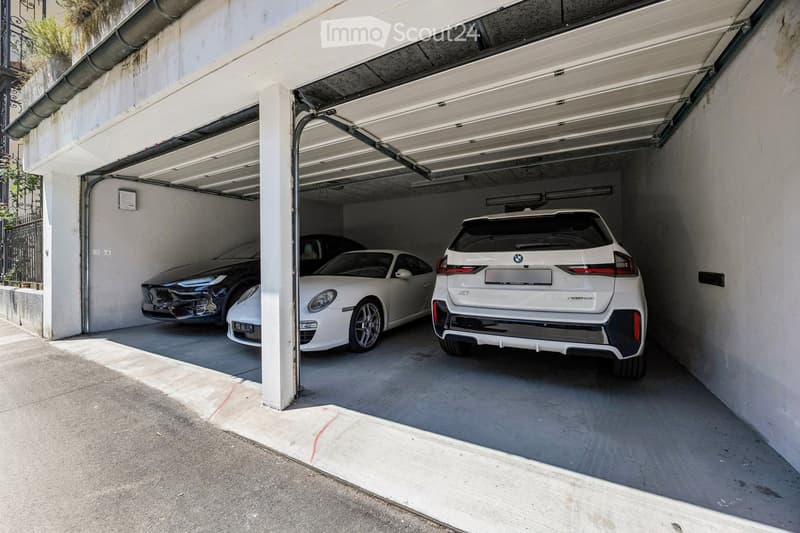 Underground parking area with three cars parked inside, including a white BMW and a white Porsche, under a concrete ceiling with fluorescent lighting.