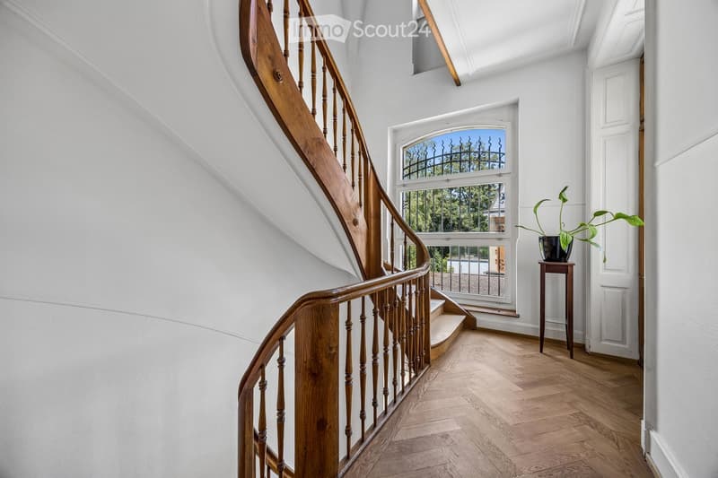 Spacious hallway with wooden flooring, white walls, and a wooden staircase with a curved handrail. A small table with a potted plant sits near the door, and a large window offers a view of the garden.