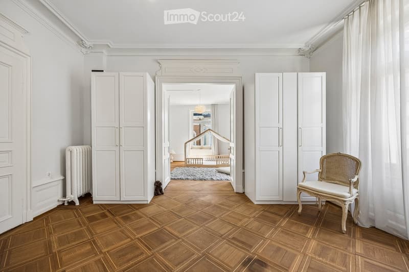Elegant hallway with wooden herringbone flooring, white walls, large built-in wardrobes, a classic white chair, and a radiator. A view into an adjacent room shows a crib and soft rug.