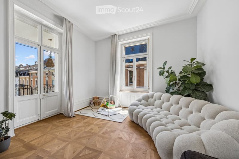 Bright living room with wooden herringbone flooring, large windows, white walls, a tufted sofa, baby stroller, toys, and potted plants.