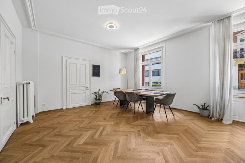 Bright dining area with herringbone wooden flooring, large window with white curtains, modern dining table with six chairs, two potted plants, radiator, and a floor lamp.