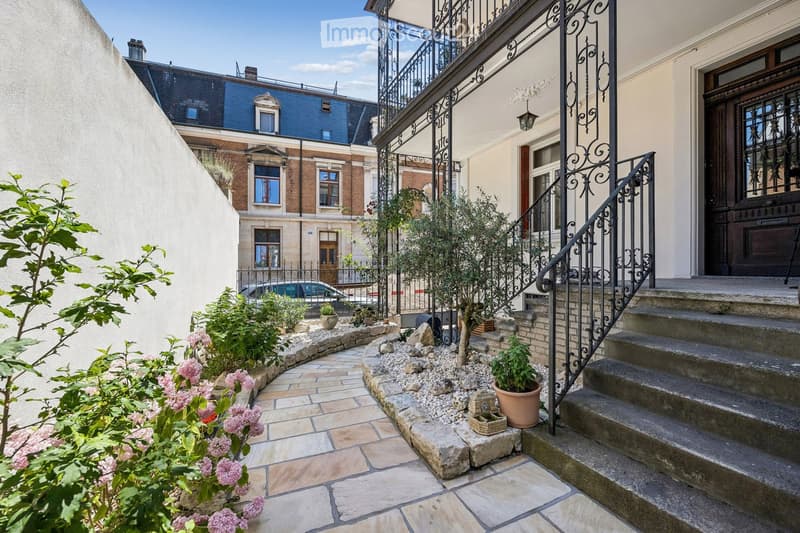 Courtyard with stone-tiled pathway, potted plants, small tree, decorative railing leading to a staircase, and surrounding buildings.