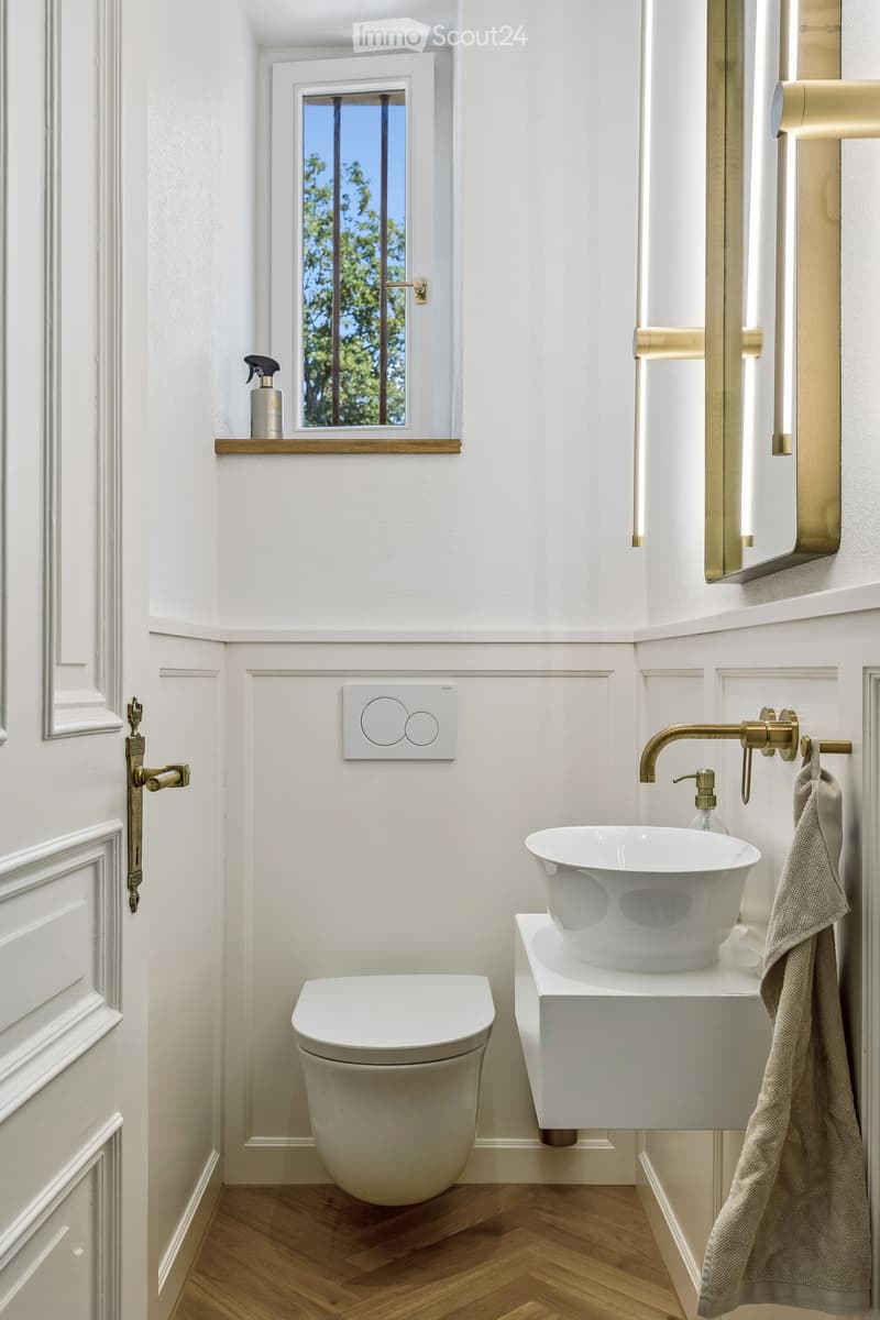 Modern bathroom with white walls, wooden floor, toilet, sink, faucet, mirror, and a towel hanging on the side.