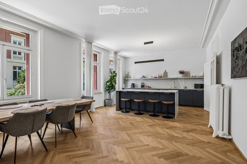 Open-plan kitchen and dining area with wooden herringbone flooring, white walls, and large windows. Features a dark blue kitchen island with bar stools, a wooden dining table with six chairs, and a microwave on the counter.