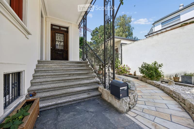 Staircase leading to a building entrance with decorative wrought iron railing, potted plants, and a mailbox on a stone platform.