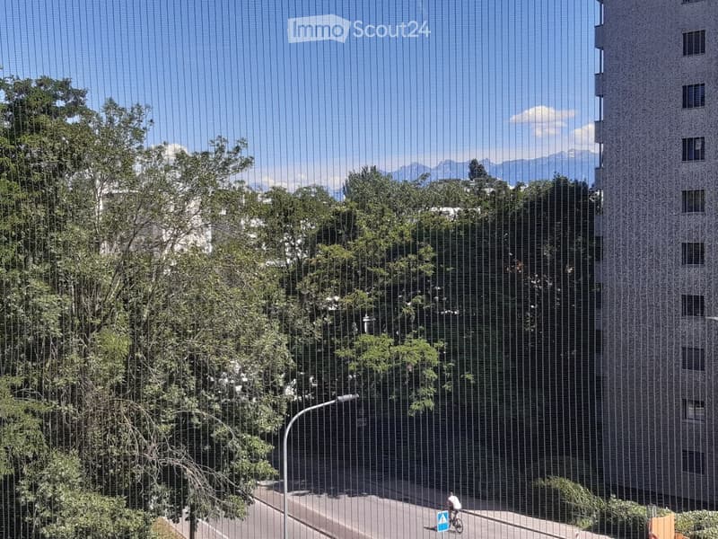 A modern multi-story building with a textured facade, surrounded by trees and a clear blue sky. The scene includes a street with a cyclist and a small blue sign on the ground.