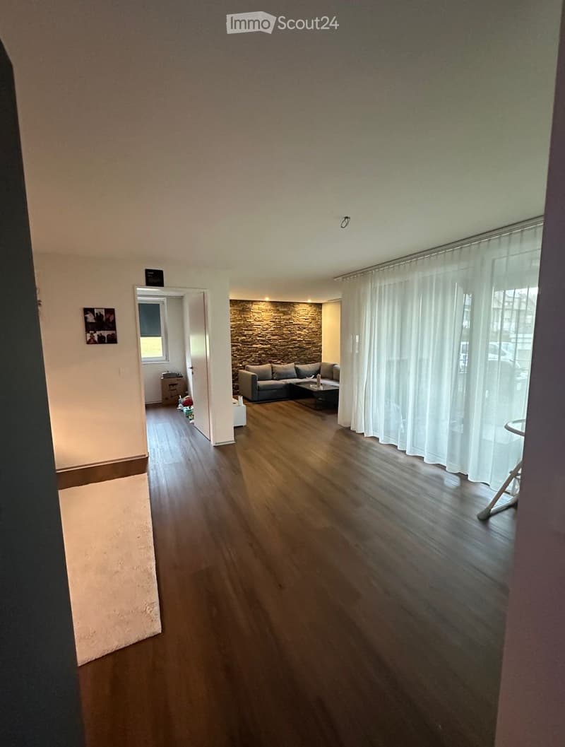 Spacious living room with wooden flooring, large windows with white curtains, a stone accent wall, and a grey sectional sofa with a coffee table.