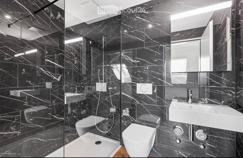 Modern bathroom with black marble tiles, two sinks, two toilets, a large mirror, and a glass-enclosed shower stall.