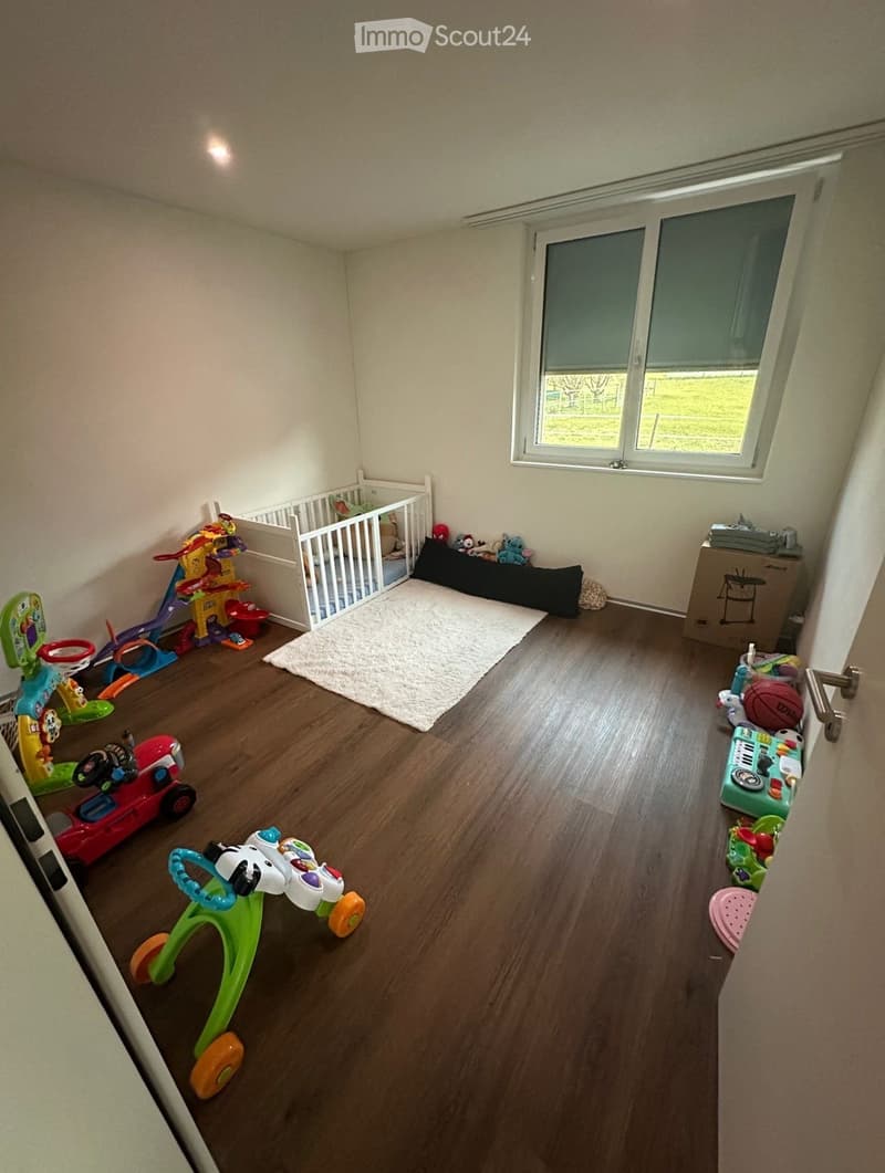 A well-lit children's room with wooden flooring, a baby crib, various toys including a toy car, basketball hoop, walker, slide, and storage boxes, and a large window with blinds.