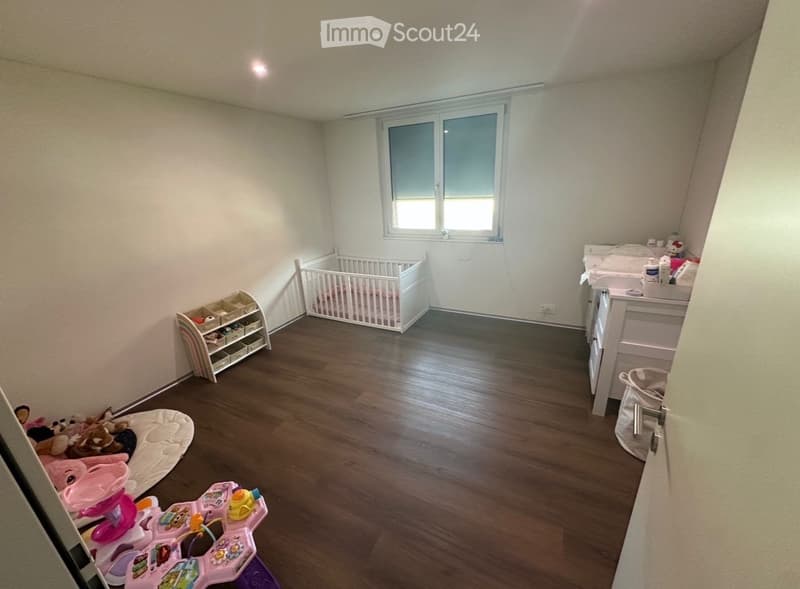 A well-lit nursery with wooden flooring, a crib, changing table, toy shelf, and various toys including a toy car and a toy chair.