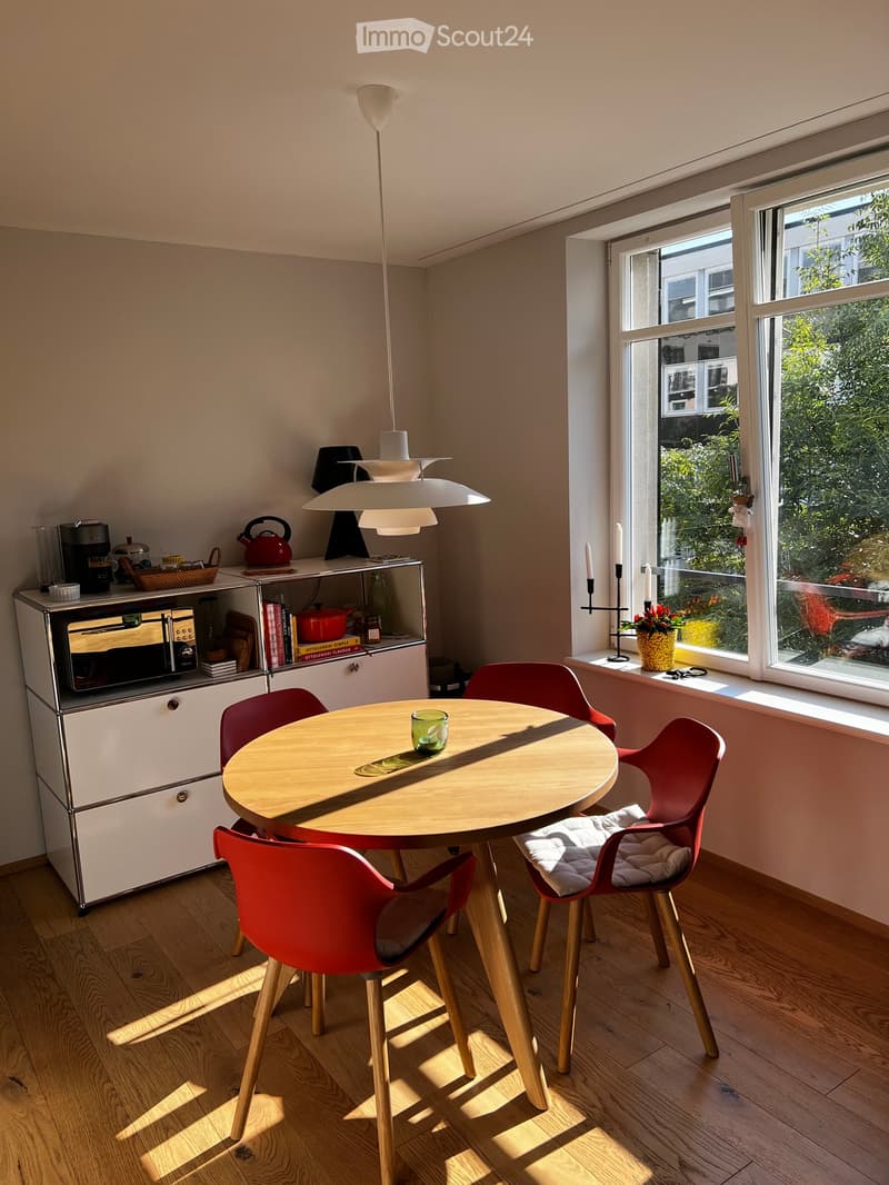Bright kitchen with a round wooden dining table surrounded by red chairs, a white cabinet with an oven, and a pendant light hanging above. A window offers a view of greenery outside, and a small flower pot sits on the windowsill.