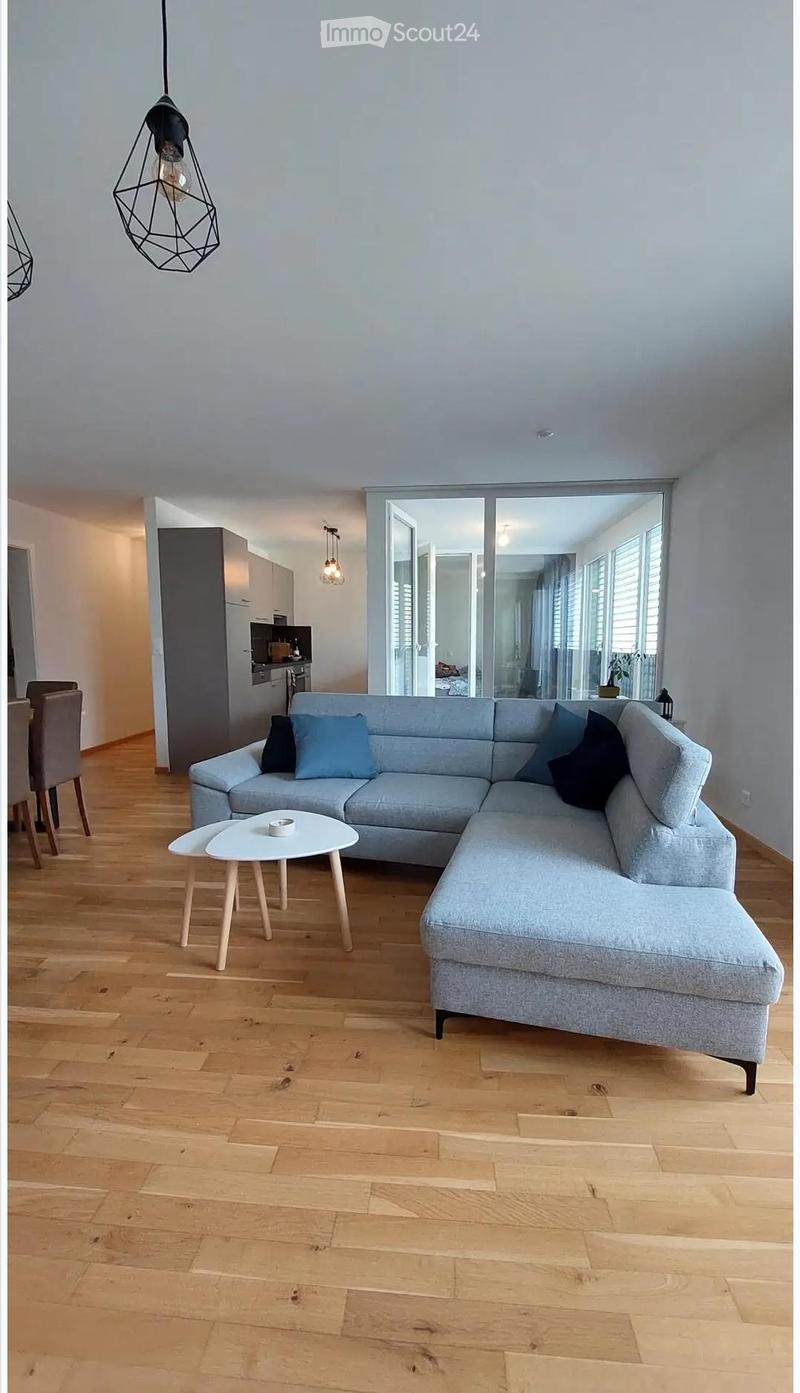 Modern living room with light grey sectional sofa, wooden coffee table, and dining area visible in the background.