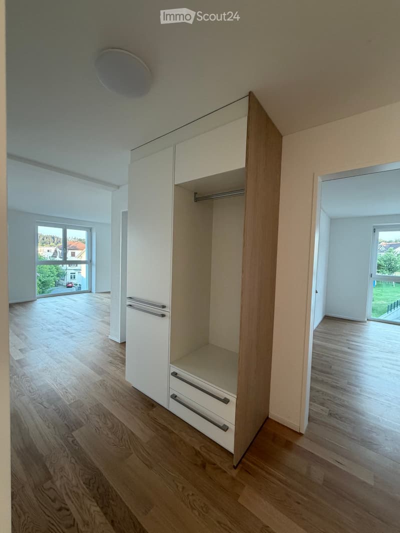 Empty room with wooden flooring, white walls, and built-in closet with drawers and shelves. Large windows provide natural light and views of the outdoors.