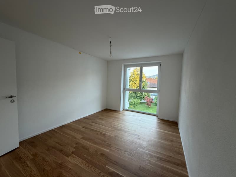 Empty room with light-colored walls, wooden flooring, and a large window showing a view of greenery outside.