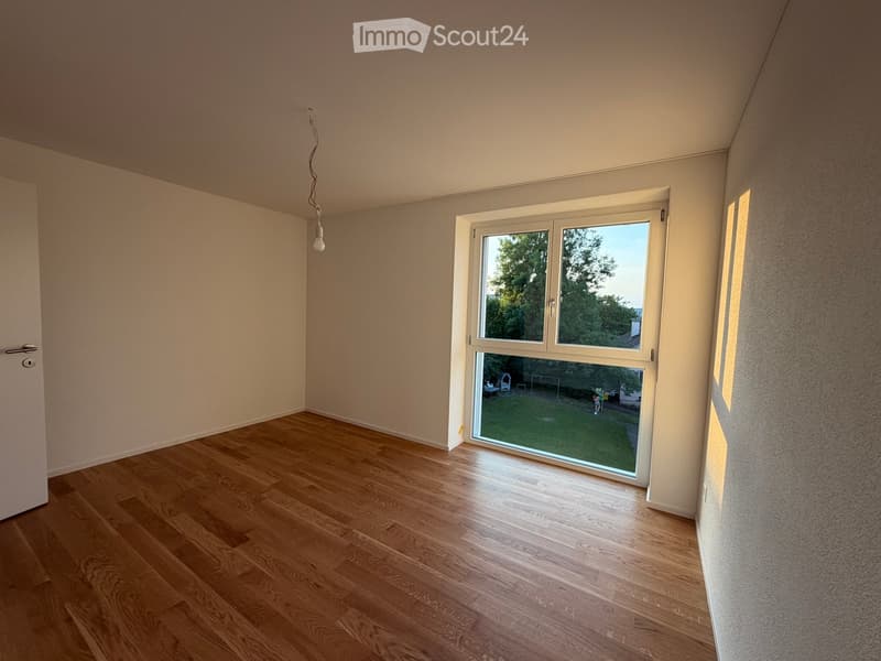 Empty room with wooden flooring, white walls, and a large window overlooking a green yard.