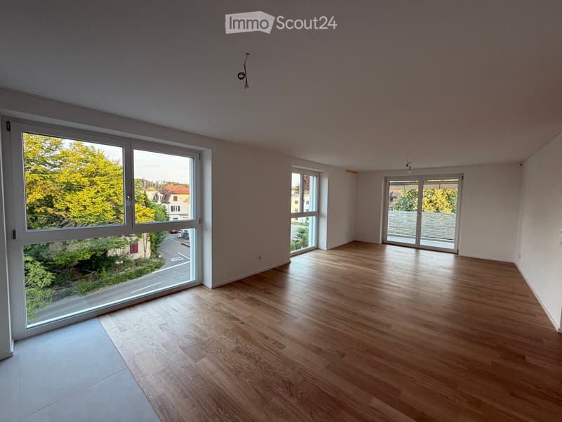 Empty room with wooden flooring, large windows, and sliding doors leading to a balcony or outdoor area.