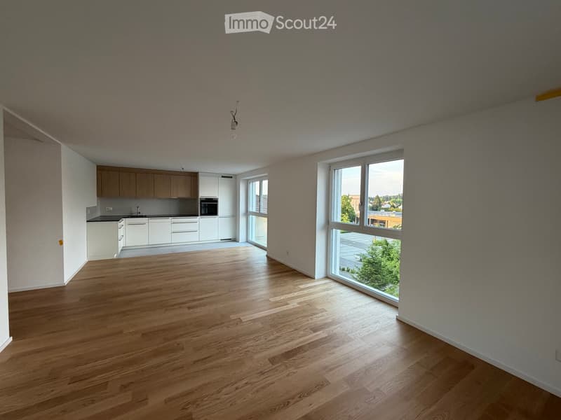 Open-plan unfurnished room with wooden flooring, large windows, and a small kitchen area with cabinets and appliances.