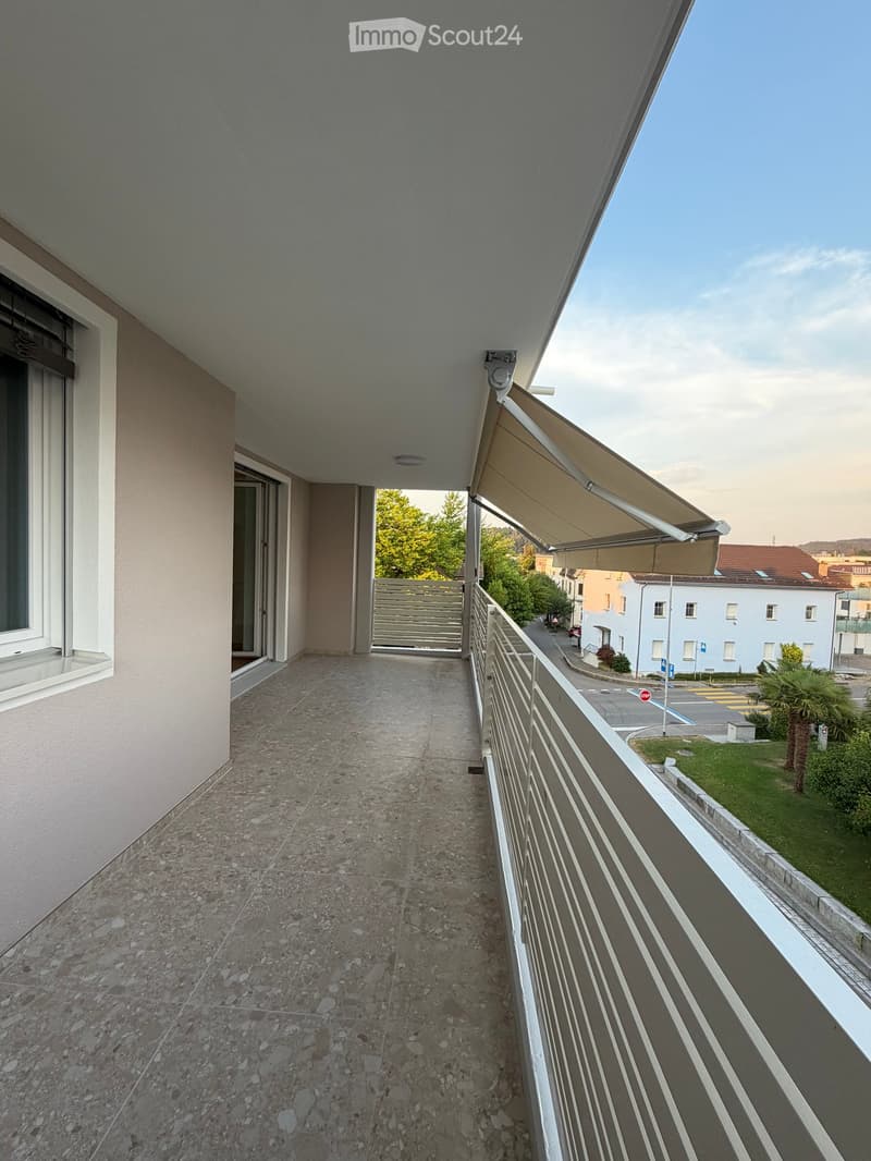 Balcony with tiled floor, metal railing, and an awning, overlooking a street with buildings and greenery.