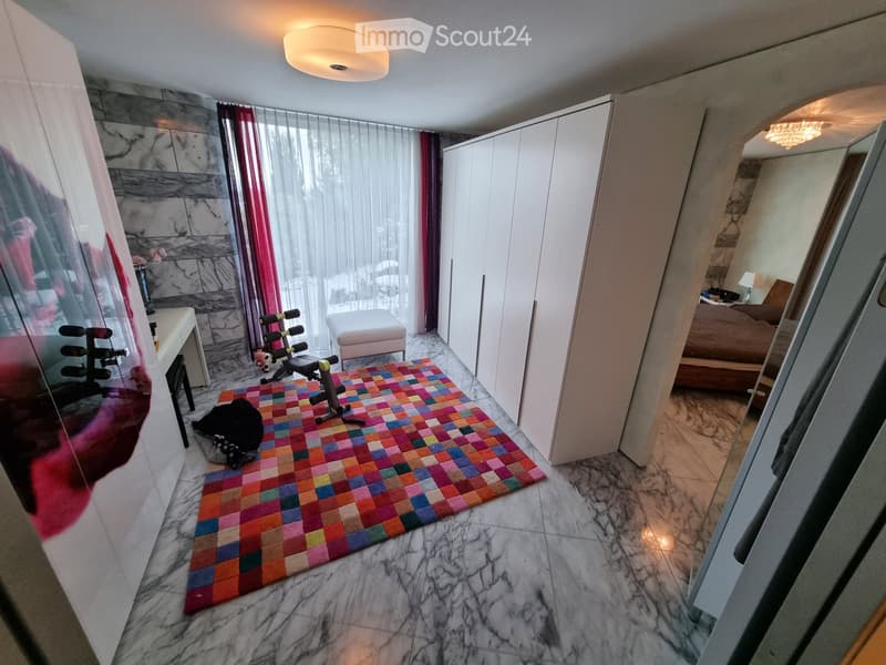 A modern, well-lit room with a colorful geometric rug, a fitness bike, a small table, a stool, a mirror, and a wardrobe. A cat is resting on the rug.