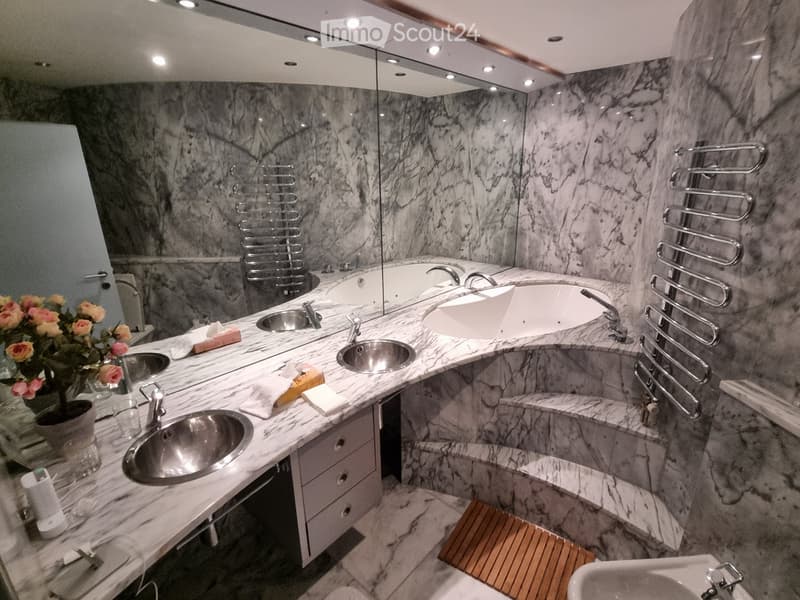 Luxurious bathroom with marble walls and floors, multiple sinks, a bathtub, towel racks, and a flower vase on the counter.
