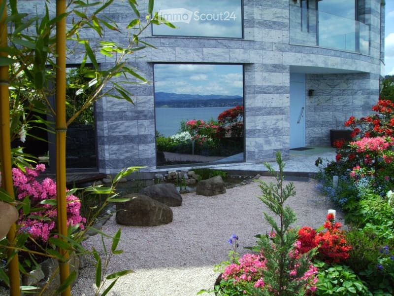 Modern house with large glass windows overlooking a scenic water view, surrounded by a well-maintained garden with colorful flowers and rocks.