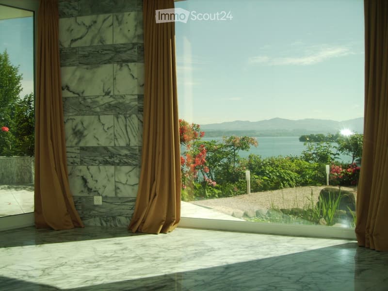 Bright room with marble walls and floor, large window offering scenic lake and mountain view, brown curtains.