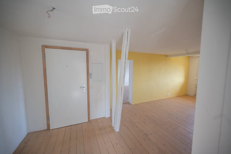 Empty room with wooden flooring, white walls, and a white door with a wooden frame. The adjacent room has yellow walls and wooden flooring.