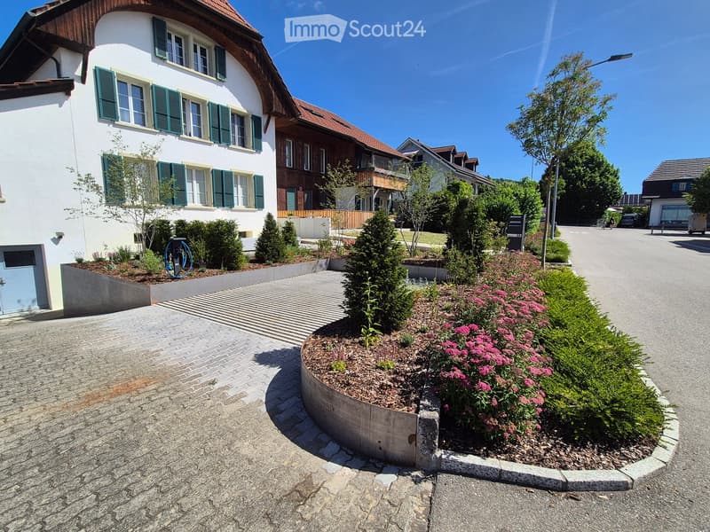 Two-story house with white walls, green shutters, and a red-tiled roof. The front features a well-maintained garden with pink flowers and shrubs, a paved driveway, and a blue electric vehicle charging station.