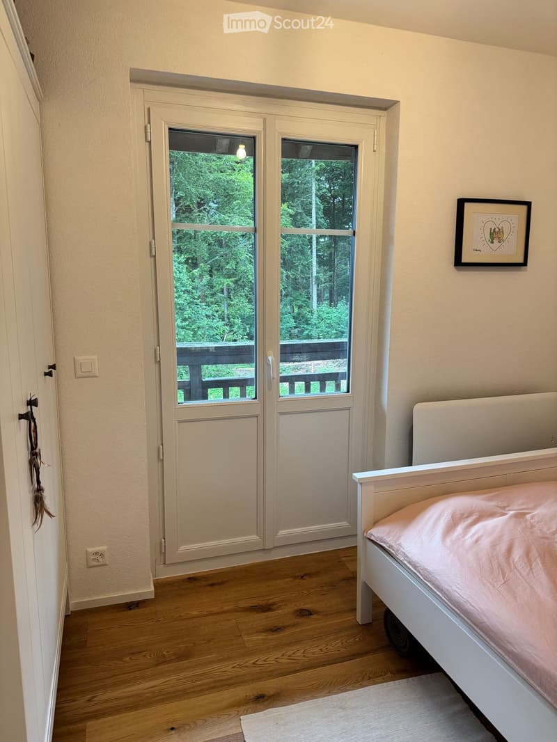 Cozy bedroom with wooden flooring, white bed with pink bedding, large window with view of greenery, and a framed picture on the wall.